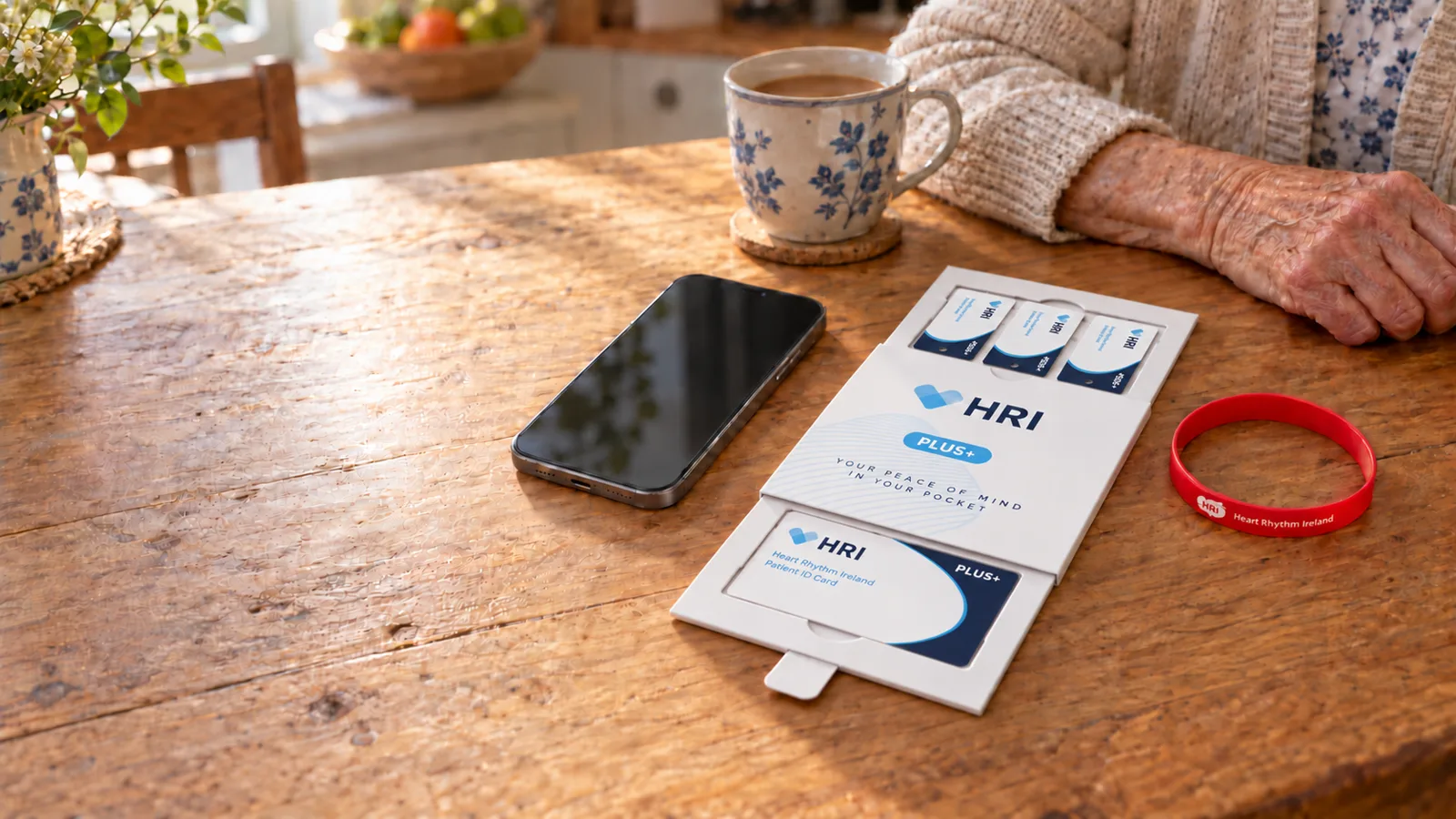 An open HRI Plus pack with a device card, three key fobs and a red wristband on a kitchen table beside an older woman's hand.
